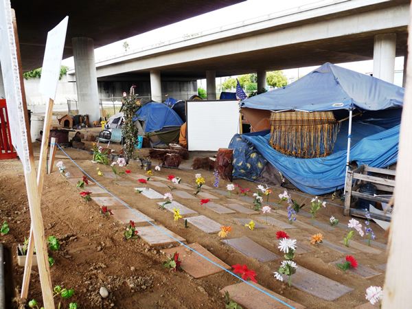 A makeshift encampment of a group of homeless people in Fresno, California, USA. The residents bordered their encampment with a homeless memorial with cardboard gravestones with the names of those who passed away on the streets marked by artificial flowers – an expression of both the dignity and resilience of this community and sad reminder of the perils faced by those without shelter. Photo by Christopher Herring.