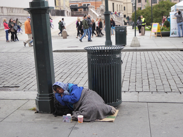 Even in Norway, with very visible government attention and programs, there are an estimated 6200 people with no place to live. Here, in the capital city of Oslo, at the steps of the Parliament Building on the busy avenue connecting the town center with the Royal Palace, a homeless woman tries to sleep and collect a few coins. (Photo by Benjamin Clavan, 2015)